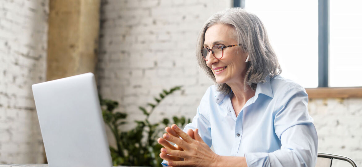 An attractive older, female patient wearing glasses and a light blue shirt sits, hands clasped and smiling looking at her laptop screen during an appointment with our Neurofeedback Therapist. She wears a light blue button up shirt. She is sitting at a desk. Behind her are white, brick walls, a bright and airy window and a eucalyptus plant in the corner.