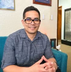 John Joseph the the patient care coordinator at Brain Health Clinic. He is pictured wearing a blue button up shjrt, and glasses. He smiles at the camera while folding his hands in front of him. He sitting in a turquoise chair next to a beige wall with a couple small pieces of artwork hanging behind him. 