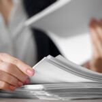 Close-up of a person’s hands handling a stack of white papers. One hand is flipping through the papers while the other holds a sheet. The person is wearing a white shirt and a dark blazer. Their nails are painted light pink.