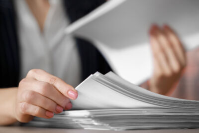Close-up of a person’s hands handling a stack of white papers. One hand is flipping through the papers while the other holds a sheet. The person is wearing a white shirt and a dark blazer. Their nails are painted light pink.