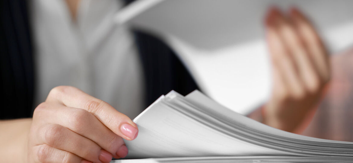 Close-up of a person’s hands handling a stack of white papers. One hand is flipping through the papers while the other holds a sheet. The person is wearing a white shirt and a dark blazer. Their nails are painted light pink.